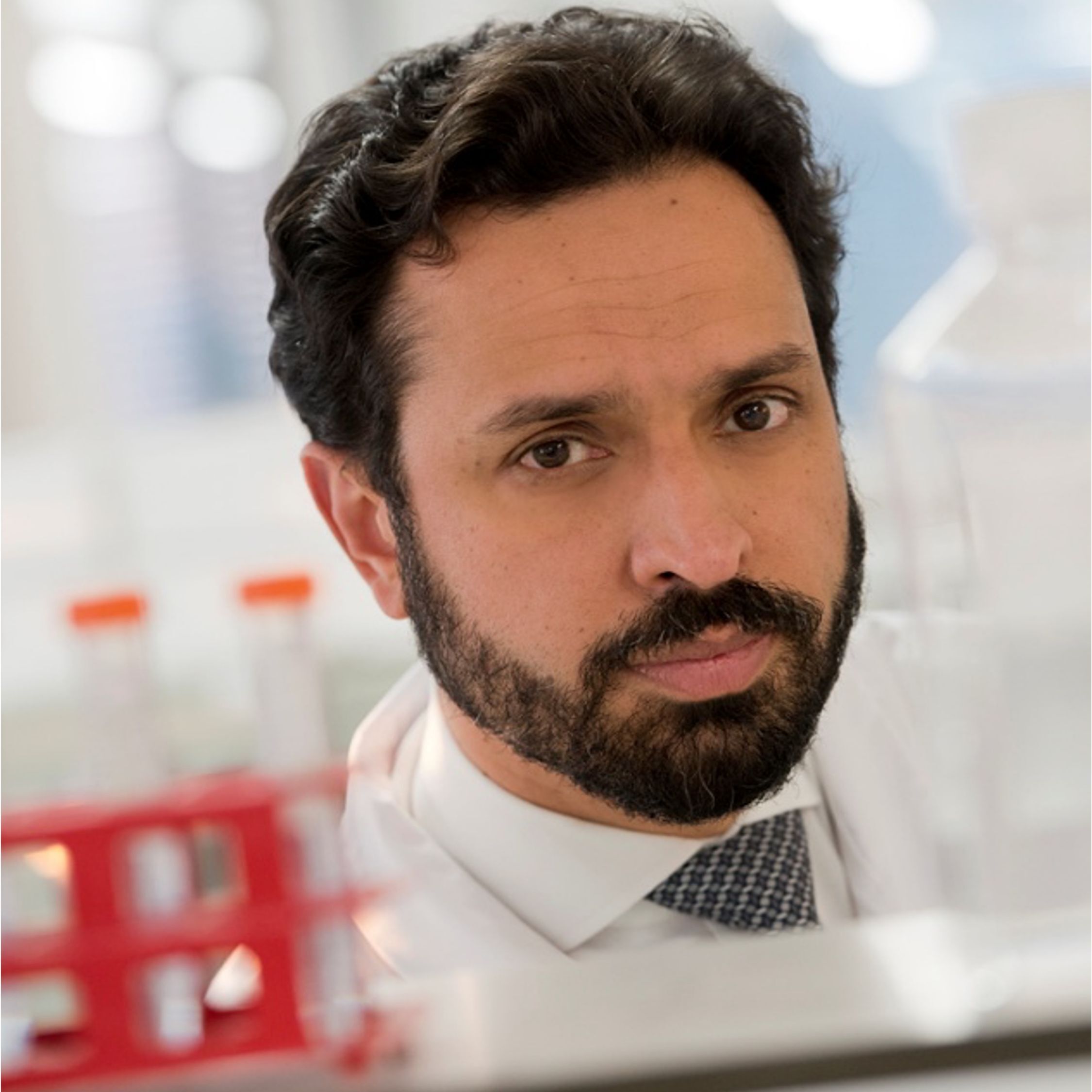 Man with dark curly hair and beard in white lab coat with tie, photographed in laboratory setting with red test tube rack visible
