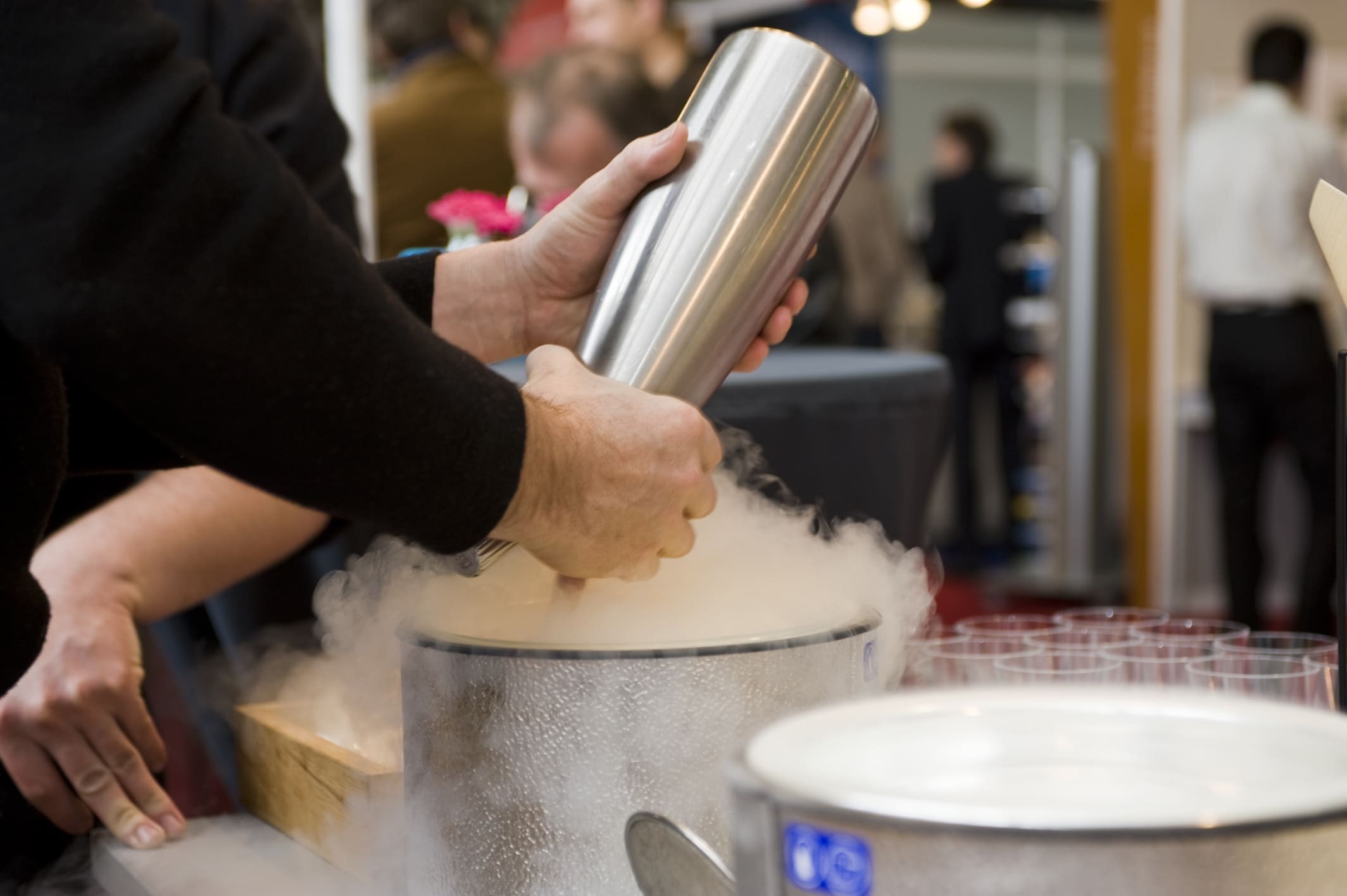 Hands preparing molecular gastronomy cocktails with liquid nitrogen vapor rising from stainless steel container at event
